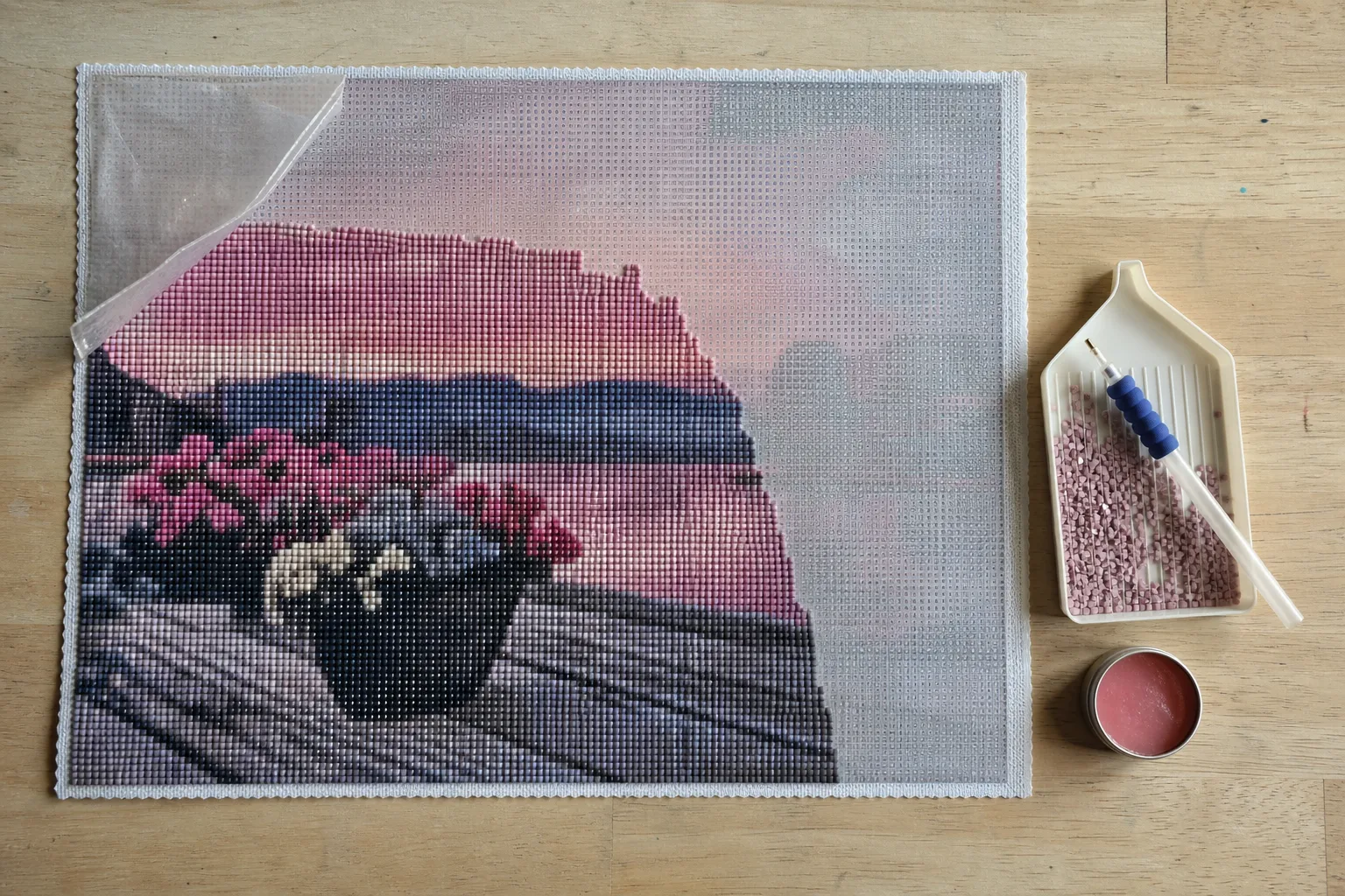 A half-finished diamond painting canvas seen from above, with the protective film peeled back at one corner, a tray of pink drills, an applicator pen and a tin of wax beside it
